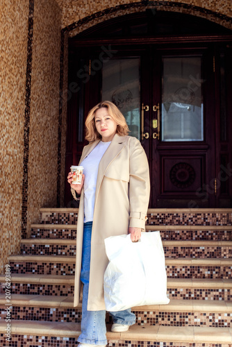 Caucasian woman in beige coat holding coffee and white shopping bag stands on steps of building with decorative tile work and wooden doors in the background