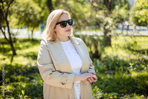 Caucasian female in sunglasses and a beige coat stands in a park surrounded by greenery, with sunlight filtering through the trees