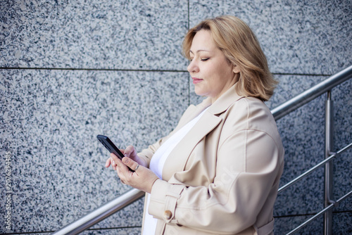 Caucasian female stands against a stone wall, using a smartphone while holding onto a metal railing, dressed in a beige coat