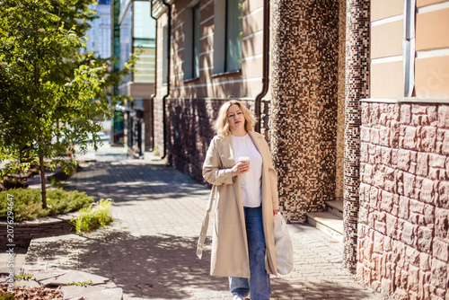 Caucasian female strolls down a city sidewalk, holding a coffee cup, surrounded by modern architecture and greenery on a sunny day