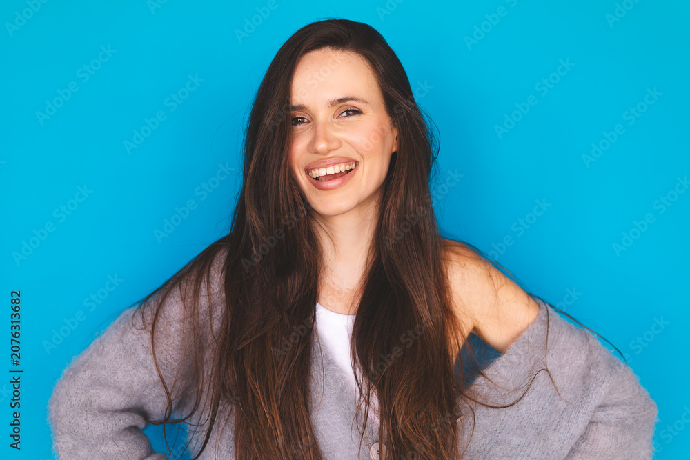 Fototapeta premium Portrait of a happy young woman with long brown hair smiling genuinely at the camera, wearing a cozy oversized grey mohair sweater, isolated on a bright blue background.