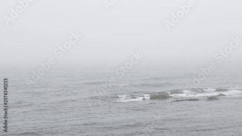 Grey stormy sea with waves crashing on coastal rocks beneath a foggy overcast sky