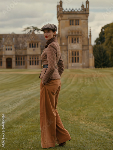 Elegant woman in vintage tweed jacket and flat cap standing in front of historic English country estate manor house