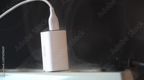 White charger emits thick smoke. USB cable plugged securely. Black background shows overheat. Charger overheats producing visible smoke. Electrical device malfunction scene. Safety warning footage