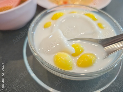 Close-up of traditional Asian dessert featuring soft milk pudding (Tao Huay) topped with yellow ginkgo nuts in sweet fresh milk, served in a clear glass bowl with a spoon
