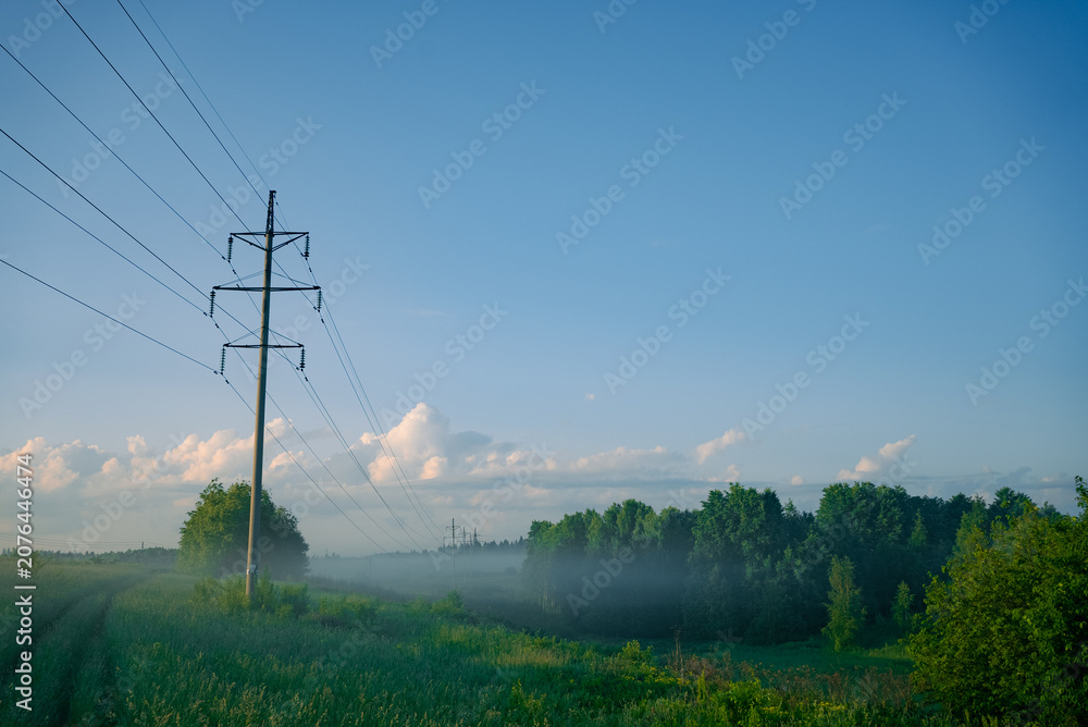 Naklejka premium Power lines extend across a vibrant green field towards a distant forest partially obscured by morning fog, showcasing utility infrastructure in a natural, open landscape under a clear blue sky