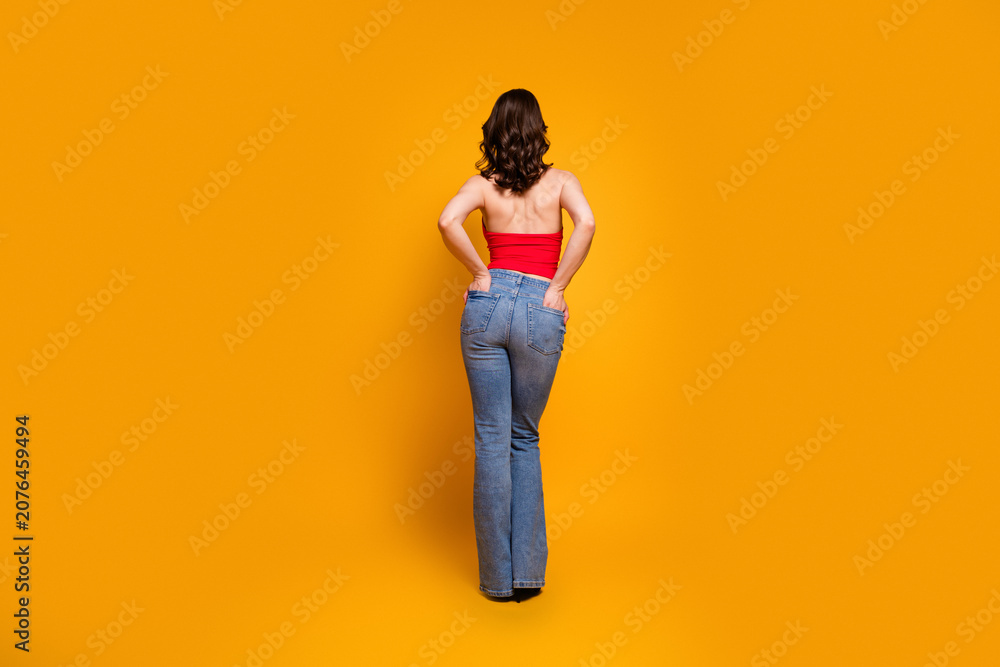 Fototapeta premium Woman standing with hands on hips wearing red top and blue jeans against vibrant orange background, rear view fashion portrait