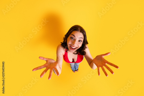 Bild auf Leinwand Happy young woman reaching up with open hands on bright yellow background, smili