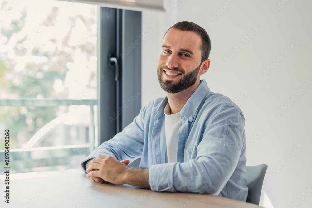 Fototapeta premium Smiling young bearded man sitting at a table near a window. Looking directly at the camera with a happy expression. Portraying positivity. Confidence. And a casual lifestyle in a bright indoor setting
