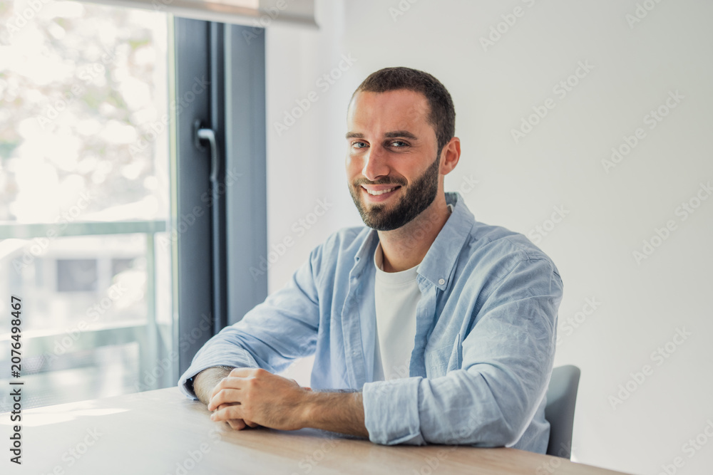 Fototapeta premium A smiling young man in a light blue shirt sits at a wooden desk by a large window, conveying relaxed confidence and approachability in a bright home office or remote work environment.