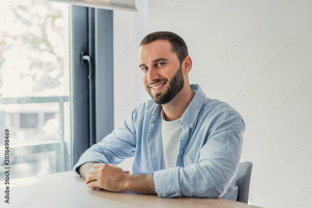 Fototapeta premium Bearded man in a blue shirt and white t-shirt sitting at a wooden table smiling at the camera. Embodying a relaxed. Happy. And confident lifestyle in a bright. Modern indoor setting with natural light