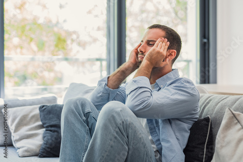 Obraz na plátně Man sitting on a sofa, suffering from a painful headache and profound stress, to