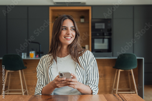 Photography A young woman sits at a kitchen table smiling while holding her smartphone, enjoying a relaxed morning at home in a modern kitchen