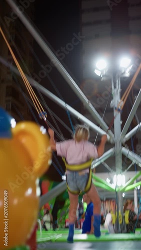 Young girl bouncing high on a bungee trampoline at an outdoor amusement park at night, secured in a safety harness with elastic cords, bright floodlights and tall buildings behind