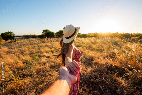 Canvas-taulu A woman in a sun hat leads her partner by the hand across a golden field at sunset, capturing a romantic travel moment full of warmth, freedom and summer adventure