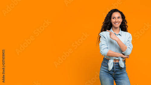A woman stands with her arms crossed and smiles while pointing to the side against a bright orange backdrop. Her curly hair and casual outfit add a relaxed vibe to the scene