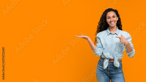 A woman with curly hair smiles while standing in front of an orange wall. She points with one hand and holds the other hand open. The setting is bright and simple, focusing on her playful pose