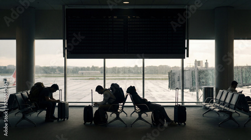 Tired passengers wait in airport lounge beside rainy runway windows with luggage flight board silhouettes delays layover fatigue business travel uncertainty disrupted trip atmosphere