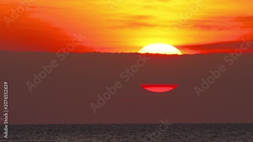 Sun rising behind dark clouds over the sea under a bright orange sky at sunset, telephoto close-up shot