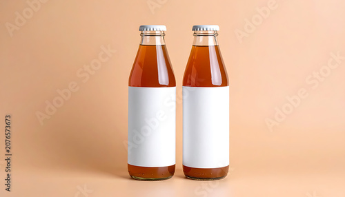 Two glass bottles with blank labels containing amber tea, isolated on beige background, branding mockup for beverage industry, organic product packaging
