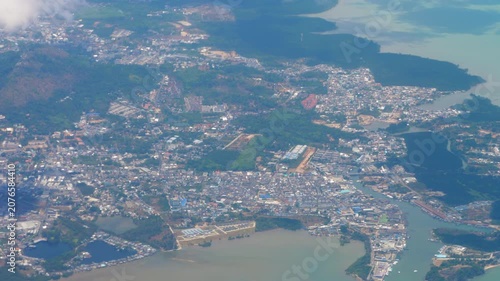 Phuket coastline and city buildings viewed from descending airplane window under bright daylight with passing clouds, high angle wide shot