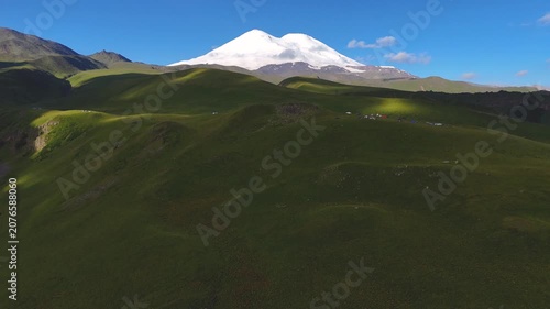 Drone view of the snow-capped peaks of Mount Elbrus and its green slopes at sunrise, with clouds in the sky