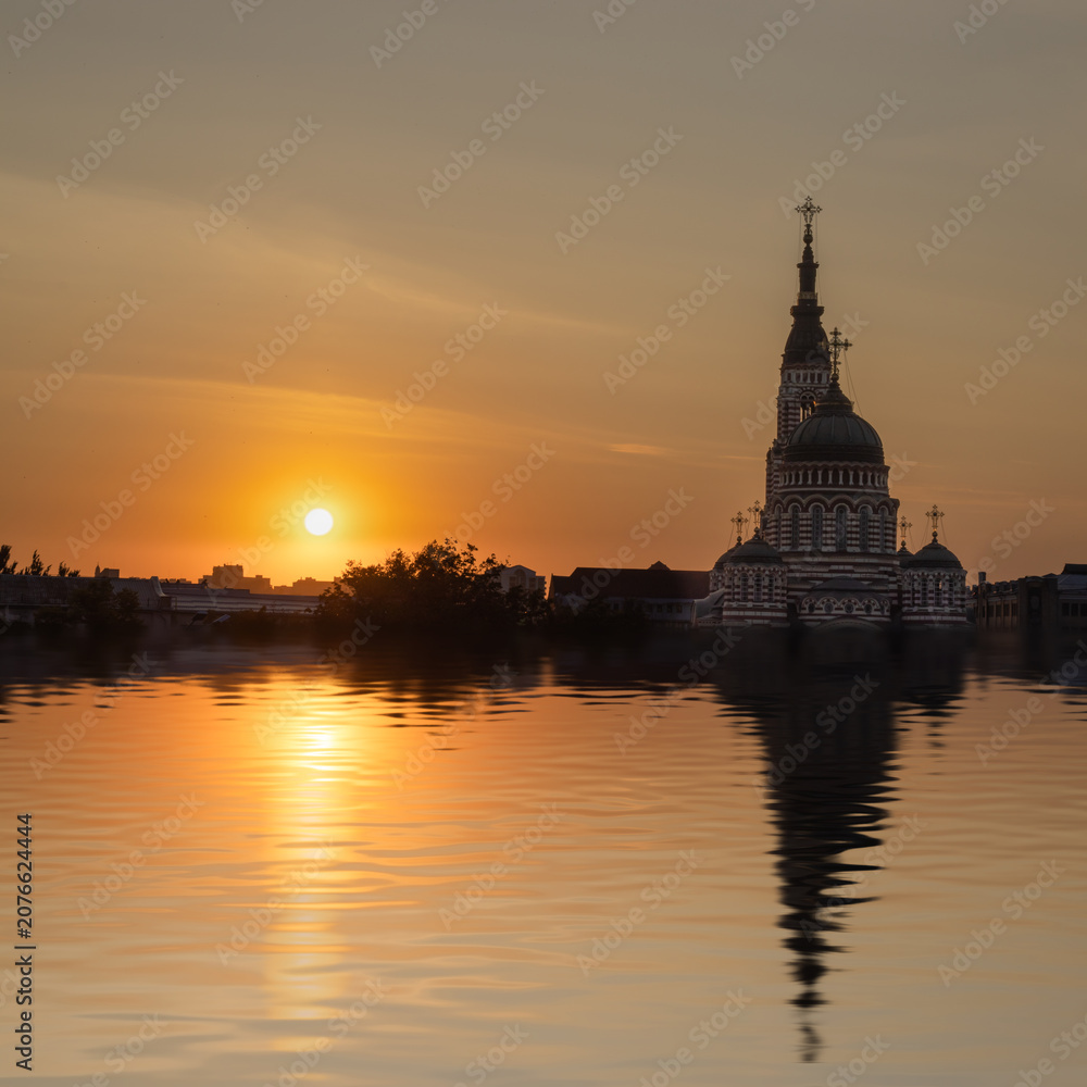 Obraz premium The sun sets behind a tall church with domes and crosses. The sky shows warm colors and the water reflects the building. It is evening near a city with a calm atmosphere.
