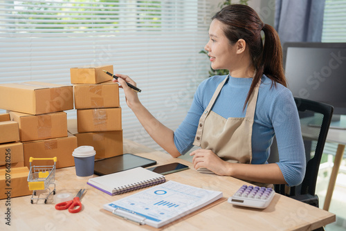 Smiling young Asian woman online seller packaging cosmetics skincare products into cardboard parcel boxes at home office, E-commerce small business owner concept.