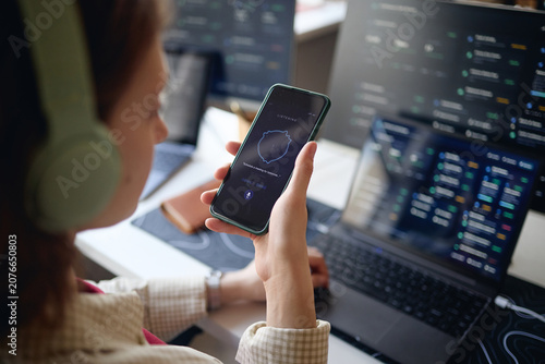 Young adult woman holding smartphone and using voice assistant while working at laptop with data dashboards, showing multitasking and AI productivity. Ideal for office work or tech marketing
