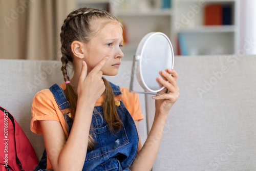 Fotografía Teen girl checking small pimple or skin blemish in handheld mirror at home, poin