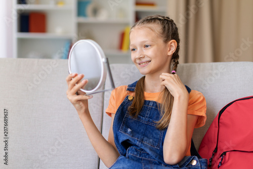 Konstfotografi Teen girl sitting on sofa at home and looking into handheld mirror while smiling