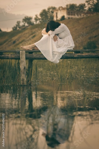 Obraz na płótnie Romantic barefooted young woman in vintage white dress sits curled up on old wooden small bridge over a calm water in summer