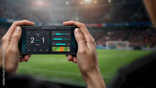 Person holding a mobile phone displaying live soccer match statistics with score updates during a professional game in a stadium