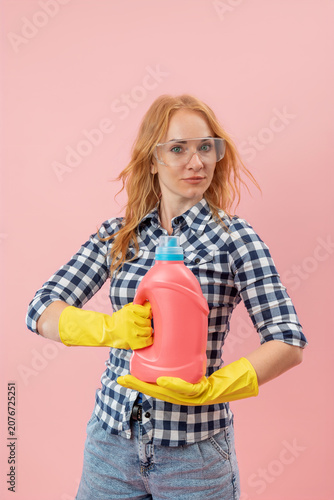 Young woman confidently holding a pink detergent bottle and wearing protective yellow gloves with safety glasses, ready for household chores, on a solid pink background