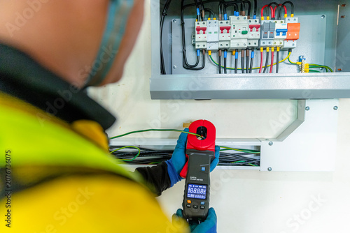 Bild auf Leinwand Electrician Testing Electrical Panel: An electrician in safety gear uses a clamp meter to test an electrical circuit breaker panel, ensuring safety and functionality