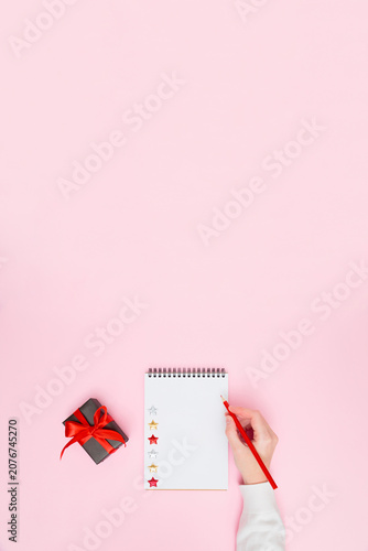 Female hand writing on blank notepad with gift box and red ribbon on pastel pink background, festive flat lay vertical banner with copy space for holiday planning, goals, to-do list or wish list.