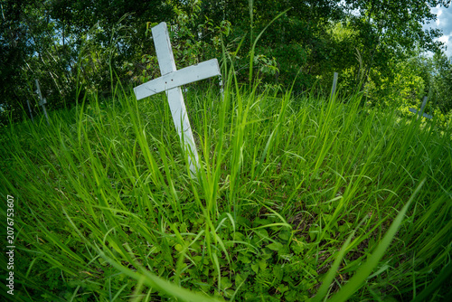 Wide-angle of an old white Christian grave marker surrounded by overgrown bright green grass in a poorly maintained cemetery.