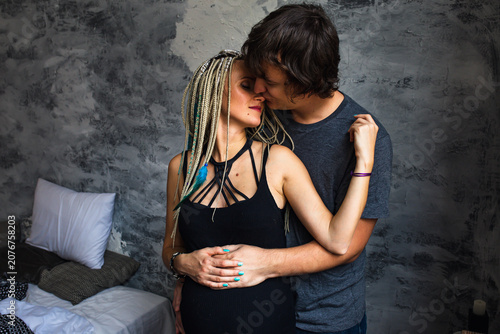 Happy pregnant woman with dreadlocks and her husband kissing near gray wall, romantic family and motherhood scene.