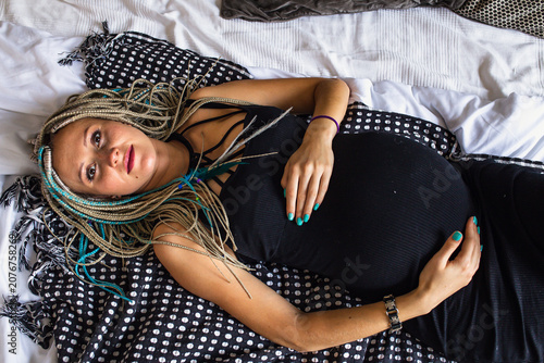 Stylish pregnant woman with dreadlocks in black dress on bed in bedroom, happy family maternity concept.