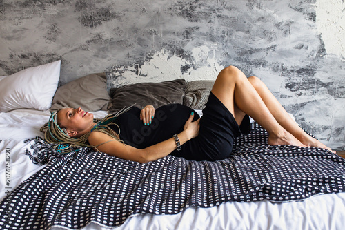 Pregnant woman with dreadlocks in black dress relaxing in bedroom, happy pregnancy and motherhood concept.