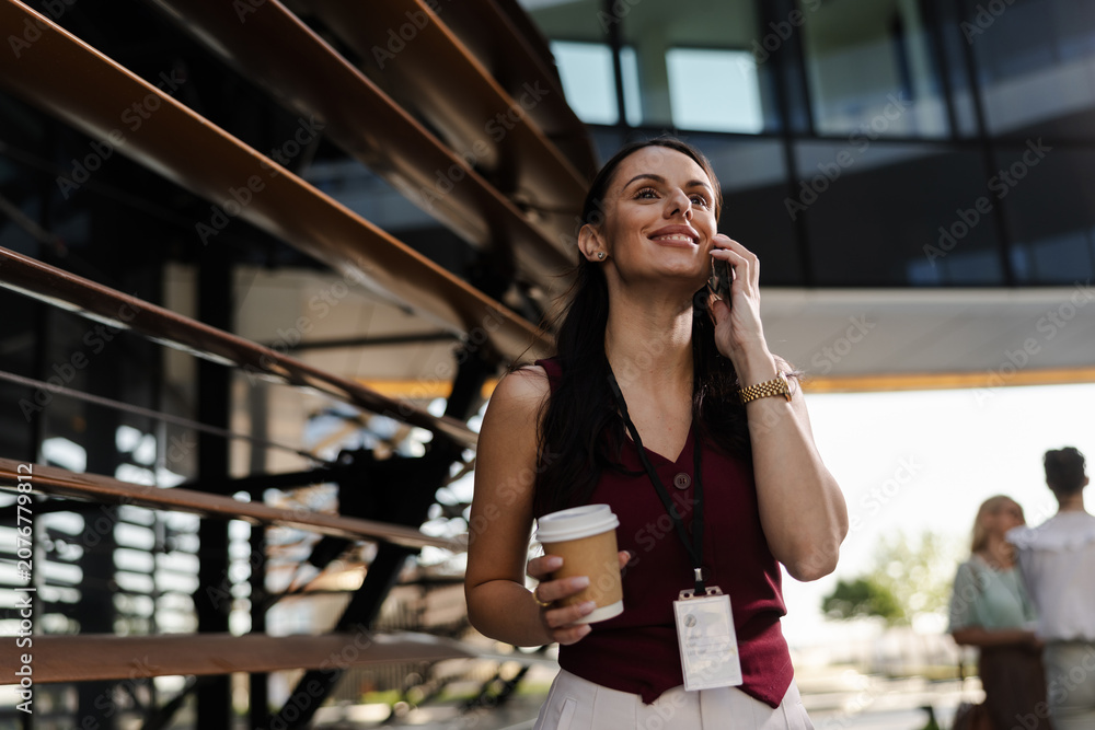 Fototapeta premium Smiling woman talking on smartphone while holding coffee cup outdoors
