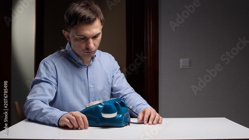 Obraz na płótnie Young man sitting at a table with an old rotary phone, patiently waiting for a c