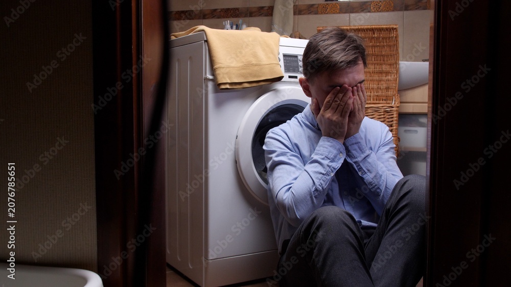 Fototapeta premium Distressed man feeling desperate, covering his face with hands, sitting alone on the floor in a laundry room, experiencing mental health issues and extreme sadness