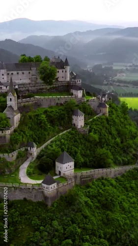 Aerial view of a historic castle complex on a lush green hill with misty mountains