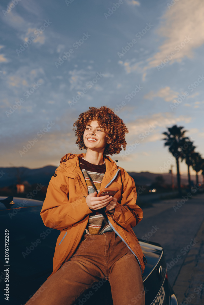 Fototapeta premium Woman lifestyle sunset outdoors portrait, smiling with curly hair, orange jacket, sitting by car on street, holding strap, warm evening light, palm trees softly blurred.