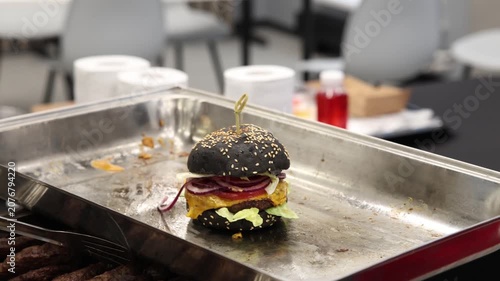 Gourmet black sesame seed burger with melted yellow cheese, red onion rings, and fresh green lettuce presented on stainless steel catering tray for indoor cooking party buffet food event.