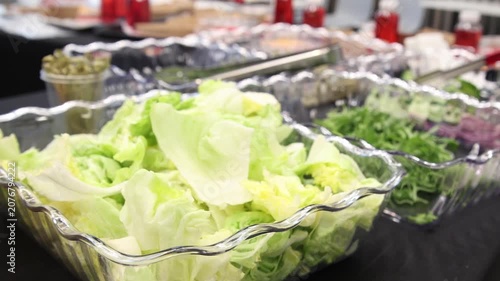 Catering buffet table featuring clear bowls with fresh green lettuce leaves, chopped herbs, and capers on a black tablecloth, with blurred red sauce bottles in the background for salad bar service.