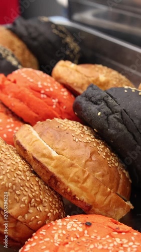 Assorted gourmet burger buns in golden, red, and black colors topped with white sesame seeds arranged in a stainless steel catering tray for a commercial event buffet service.