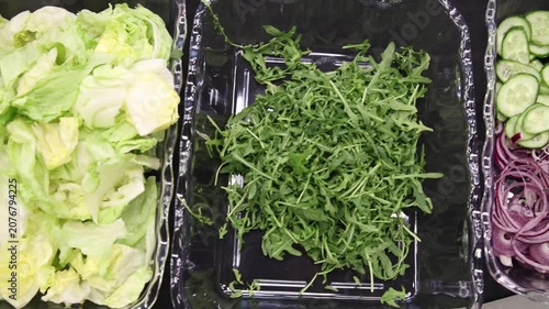 Top view of catering salad bar ingredients featuring fresh green lettuce leaves in a glass bowl, chopped herb greens in a black container, and sliced red onion rings on a black table.