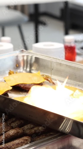 Professional chef using a bright cooking torch to melt yellow cheese slices over grilled burger patties in a stainless steel catering tray for an outdoor party buffet food preparation.